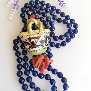 May include: A blue gemstone necklace with a ceramic pendant shaped like a basket. The basket is decorated with floral patterns in pink, green, and black. The pendant is accented with red coral.