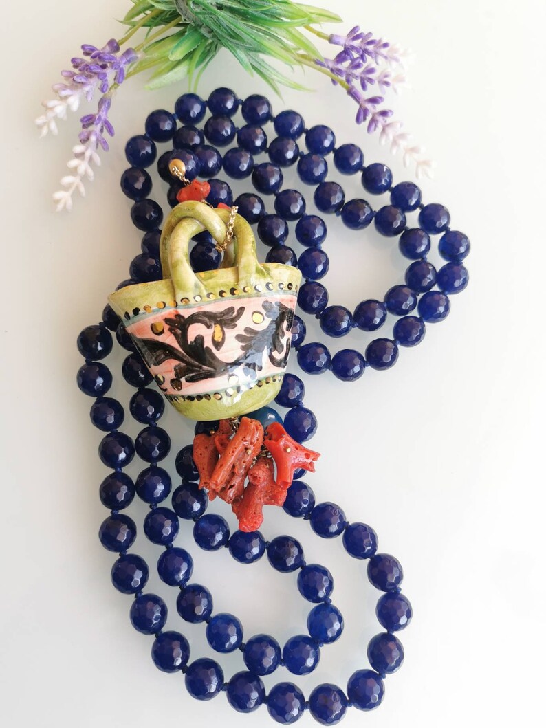 May include: A blue gemstone necklace with a ceramic pendant shaped like a basket. The basket is decorated with floral patterns in pink, green, and black. The pendant is accented with red coral.