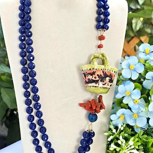 May include: A double strand necklace made of dark blue beads with a ceramic pendant shaped like a basket with floral designs. The pendant is attached to a red coral piece and a blue agate bead.