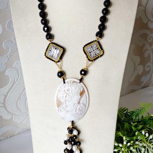 Op de afbeelding: Een elegante ketting met een camee hanger met een vrouwenprofiel. De ketting heeft zwarte kralen strengen, vierkante decoratieve accenten en een kwast van zwarte en witte kralen. De ovale camee heeft een crèmekleur.