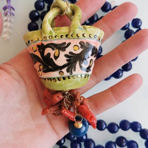 May include: A green ceramic basket pendant necklace with a gold chain and coral accents. The basket has a pink and black floral design with gold accents. The necklace is on a blue beaded chain.