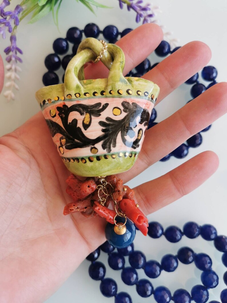 May include: A green ceramic basket pendant necklace with a gold chain and coral accents. The basket has a pink and black floral design with gold accents. The necklace is on a blue beaded chain.