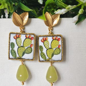 May include: Gold tone earrings with a square tile featuring a green cactus with red fruit. The tile is surrounded by a gold trim. The earrings have a green teardrop-shaped stone hanging from the bottom.