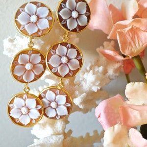 Könnte beinhalten: Ein Paar goldfarbene Ohrringe mit drei runden, geschnitzten Kameen-Anhängern mit weißen Blumendesigns. Die Anhänger sind durch goldfarbene Ketten verbunden.