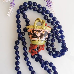 May include: A necklace made of dark blue beads with a ceramic pendant shaped like a basket. The basket is decorated with floral patterns in pink, green, and black. The pendant is accented with coral-colored beads.