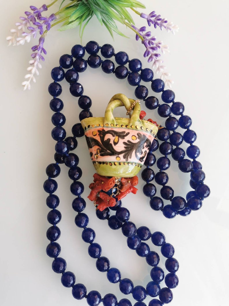May include: A necklace made of dark blue beads with a ceramic pendant shaped like a basket. The basket is decorated with floral patterns in pink, green, and black. The pendant is accented with coral-colored beads.