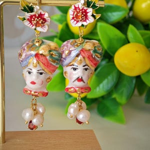 Peut inclure: Une paire de boucles d'oreilles ornées avec des visages en céramique peints à la main avec des turbans, des lèvres rouges et des perles. Les boucles d'oreilles sont surmontées de motifs floraux rouges, blancs et jaunes, et sont suspendues à des crochets dorés.
