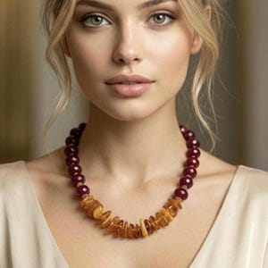 Op de afbeelding: Een ketting bestaande uit een combinatie van gladde, ronde, diepbordeauxrode kralen en onregelmatige, doorschijnende, goudbruine barnsteenkleurige schilfers. De kralen zijn in een cirkelvorm rond de nek gerangschikt, met de barnsteenschilfers aan de voorzijde.
