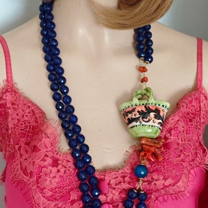May include: A double strand necklace made of dark blue beads with a green and orange ceramic pendant hanging from it. The pendant is shaped like a basket with floral designs. The necklace is worn by a mannequin with long brown hair.