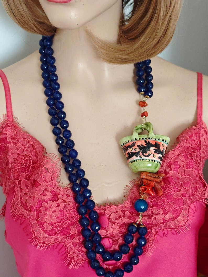 May include: A double strand necklace made of dark blue beads with a green and orange ceramic pendant hanging from it. The pendant is shaped like a basket with floral designs. The necklace is worn by a mannequin with long brown hair.