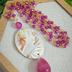 Puede incluir: Un collar dorado con un colgante de piedra rosa y un colgante de camafeo. El collar presenta múltiples hilos de pequeñas cuentas rosas y una gran gema rosa en forma de lágrima. El colgante de camafeo ovalado representa el perfil de una mujer.