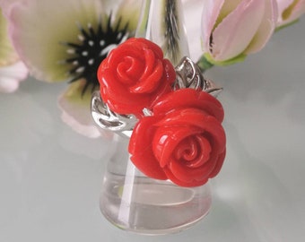 May include: A silver ring featuring two vibrant red rose-shaped adornments. The roses are detailed with intricate petal designs. The ring is displayed on a clear, conical stand, with soft-focus flowers in the background.