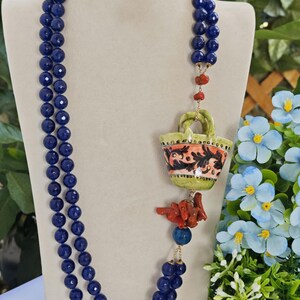 May include: A long, double-strand necklace made of dark blue beads with a ceramic pendant shaped like a basket with floral designs. The pendant is attached to a gold chain with a coral piece hanging below.