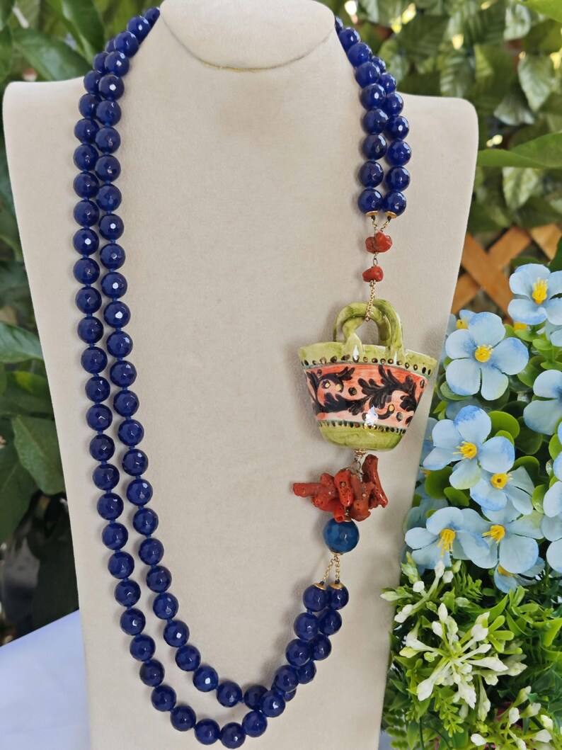 May include: A long, double-strand necklace made of dark blue beads with a ceramic pendant shaped like a basket with floral designs. The pendant is attached to a gold chain with a coral piece hanging below.