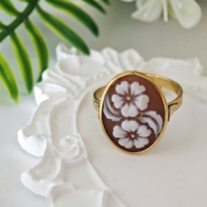 May include: A gold ring with a brown cameo featuring white carved flowers. The cameo is oval shaped and set in a gold band.