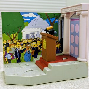 May include: A plastic playset depicting the Springfield Town Hall with a brown podium, microphones, and a red carpet. The background features a printed scene of a crowd of cartoon characters and a building. The structure includes columns and text.