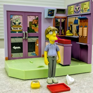 May include: A plastic playset of a fast-food restaurant interior featuring a yellow-skinned character figure with brown hair, wearing a light purple shirt and gray pants. The set includes a red tray, a yellow burger box, and a white cup accessory.