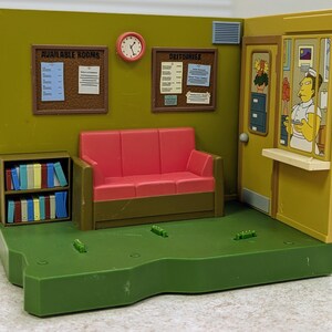 May include: A plastic playset depicting a hospital waiting room with olive green walls, a red sofa, and a small bookshelf filled with colorful books. Two cork boards labeled Available Rooms and Obituaries hang on the wall with a round clock between them.