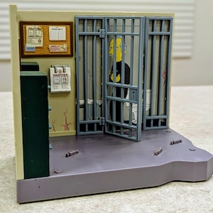 May include: A plastic playset depicting a jail cell with gray bars, a hinged door, and a beige wall featuring a corkboard with a Wanted poster and a softball sign-up sheet. A printed image of a character in a black shirt is visible behind the bars.