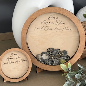 May include: Two wooden coin jars with a clear glass front. The larger jar has a stand and the smaller jar has a small stand. Both jars have the inscription "Dimes Appear When Loved Ones Are Near".