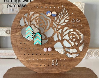 May include: Wooden earring stand with a floral design. The stand has a round base and a circular top with holes for earrings. There are several pairs of earrings on the stand, including turquoise and white earrings. The text "FREE pair of earrings with purchase." is visible at the top of the image.