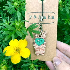 Puede incluir: Un amuleto de madera con un personaje de dibujos animados que sostiene una hoja verde. El amuleto está unido a una correa de teléfono negra con un cierre plateado. El amuleto está en una etiqueta marrón con el texto "ya haha" impreso en ella.