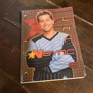 May include: Spiral-bound notebook featuring a portrait of a smiling man. The cover has the text "Lance" and "N Sync" with a star graphic. The background is brown with a red star design. The notebook is open.