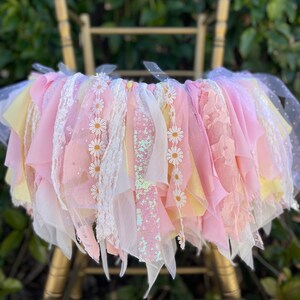 May include: A pink and yellow fabric garland with white daisy accents and a sparkly pink ribbon. The garland is hanging from a gold chair.