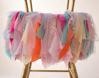 May include: A multi-colored tulle skirt draped over a gold-toned chair. The skirt features layers of pink, orange, blue, and yellow fabric strips, some with sequins and small pom-poms, creating a textured, voluminous appearance with varied lengths.
