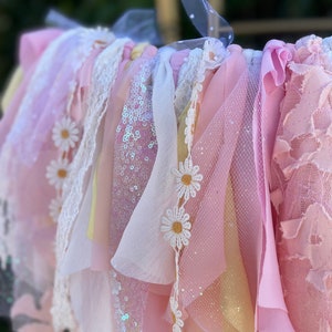 May include: A pink, white, and yellow fabric garland with daisy embellishments. The garland is made of various textures, including lace, tulle, and sequins.