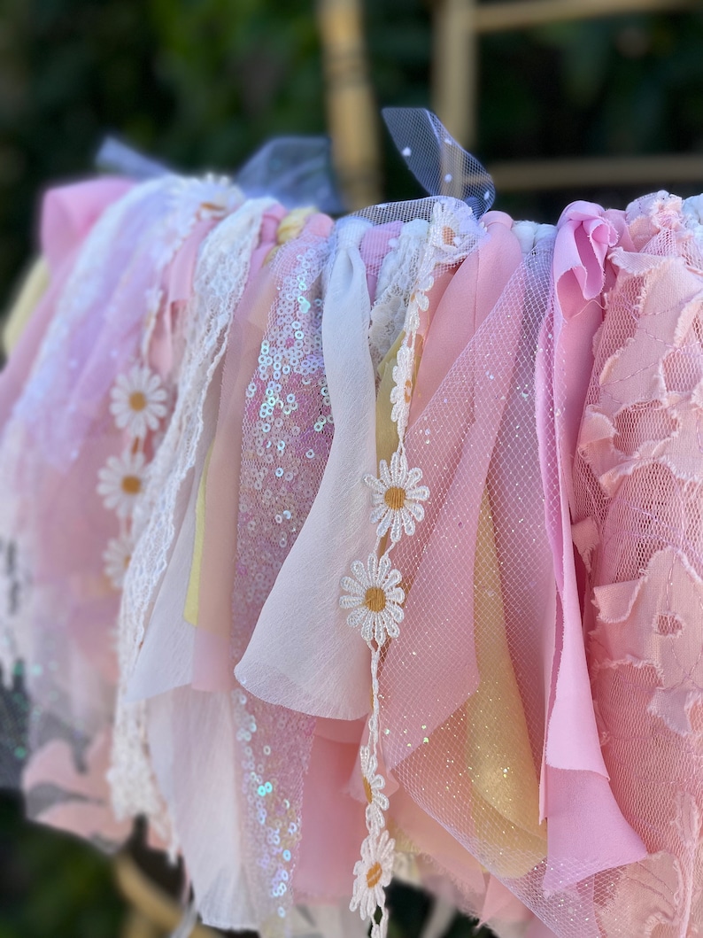 May include: A pink, white, and yellow fabric garland with daisy embellishments. The garland is made of various textures, including lace, tulle, and sequins.