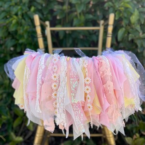 May include: A pink and yellow tulle and lace chair sash with white daisy embellishments. The sash is tied around a gold chair.