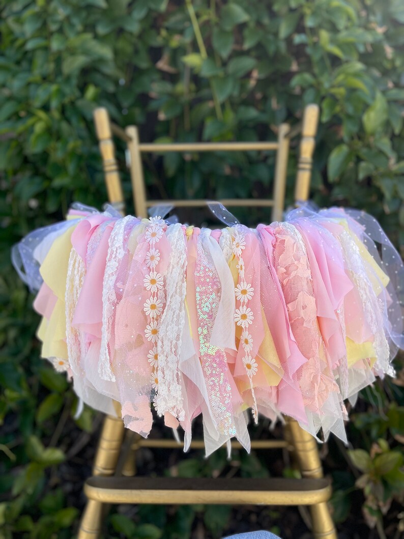 May include: A pink and yellow tulle and lace chair sash with white daisy embellishments. The sash is tied around a gold chair.