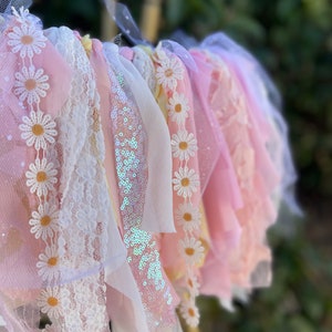 May include: A pink and white fabric garland with daisy-shaped embellishments and sequins. The garland is made of tulle, lace, and ribbon.