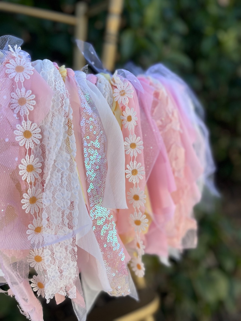May include: A pink and white fabric garland with daisy-shaped embellishments and sequins. The garland is made of tulle, lace, and ribbon.
