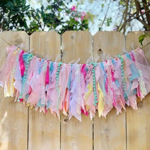 May include: A pink, yellow, and blue fabric garland with turquoise pom-poms hanging from a wooden fence.