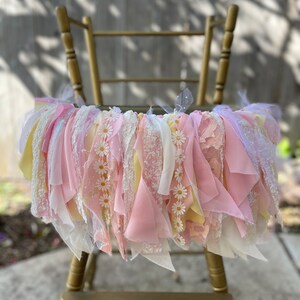 May include: A pink and yellow fabric garland with white daisy flowers, hanging on a gold high chair. The garland is made of various textures, including lace, tulle, and ribbon.