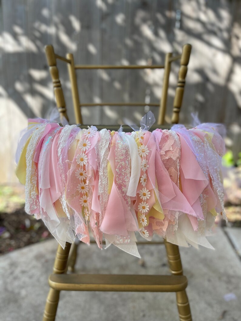 May include: A pink and yellow fabric garland with white daisy flowers, hanging on a gold high chair. The garland is made of various textures, including lace, tulle, and ribbon.
