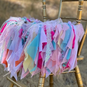 May include: A colorful tulle and fabric garland with pink, blue, yellow, and orange fabric strips, and white tulle with silver glitter accents. The garland is draped over a gold chair.