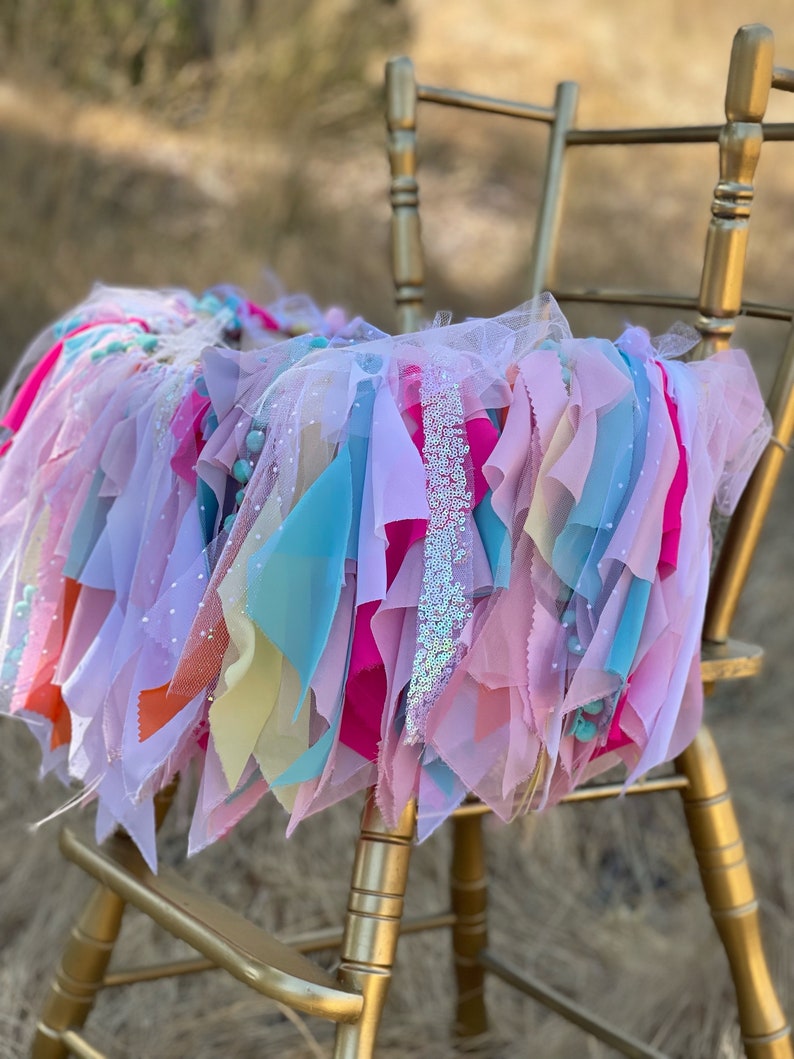 May include: A colorful tulle and fabric garland with pink, blue, yellow, and orange fabric strips, and white tulle with silver glitter accents. The garland is draped over a gold chair.