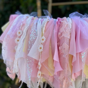 May include: A pink and yellow fabric garland with white daisy accents. The garland is made of various textures, including lace, tulle, and sequins.