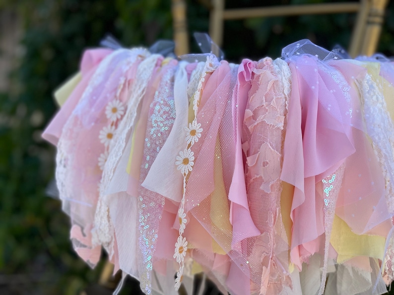 May include: A pink and yellow fabric garland with white daisy accents. The garland is made of various textures, including lace, tulle, and sequins.