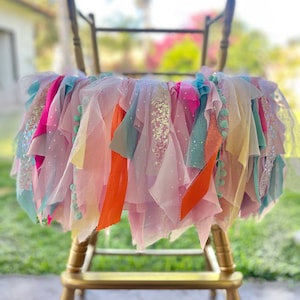 May include: A chair decorated with a colorful tulle and ribbon garland. The garland is made of pink, blue, yellow, orange, and green tulle and ribbon. The chair is gold and has a simple design.