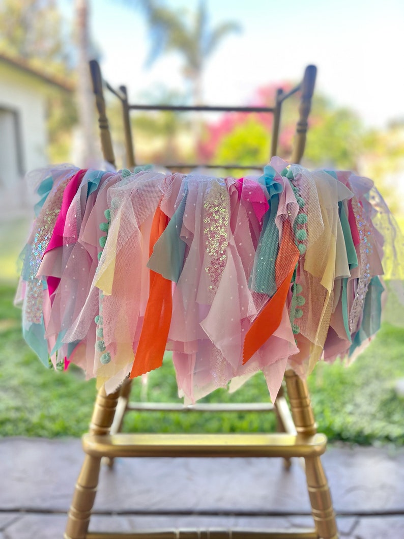 May include: A chair decorated with a colorful tulle and ribbon garland. The garland is made of pink, blue, yellow, orange, and green tulle and ribbon. The chair is gold and has a simple design.