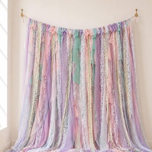 May include: A decorative backdrop made of hanging fabric strips in pastel shades of lavender, mint green, and soft pink. The curtain features alternating textures of sheer chiffon, intricate white lace, and shimmering gold sequin ribbon.