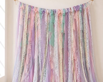 May include: A decorative backdrop made of hanging fabric strips in pastel shades of lavender, mint green, and soft pink. The curtain features alternating textures of sheer chiffon, intricate white lace, and shimmering gold sequin ribbon.