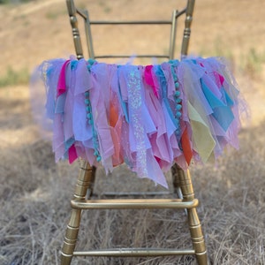 May include: A gold chair with a colorful fabric garland draped over the back. The garland is made of pink, blue, yellow, and orange fabric strips with sequins and pom-poms.