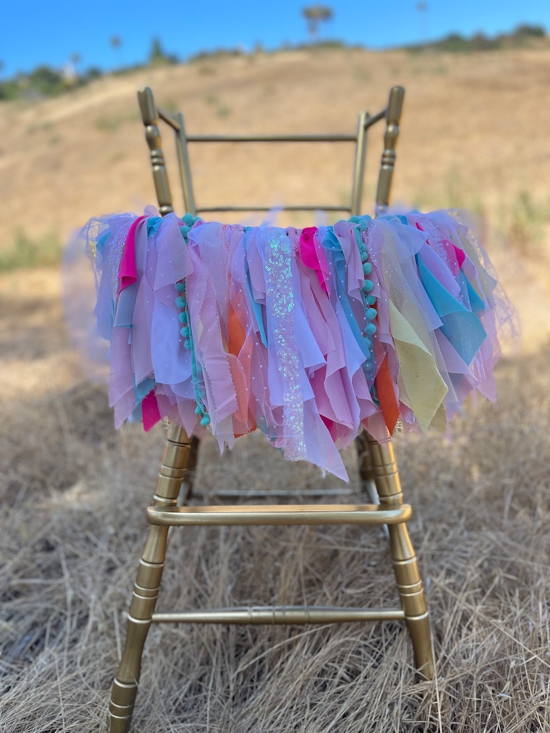 May include: A gold chair with a colorful fabric garland draped over the back. The garland is made of pink, blue, yellow, and orange fabric strips with sequins and pom-poms.