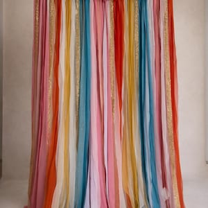 Pode incluir: Um pano de fundo decorativo feito de longas tiras verticais de tecido em tons de rosa, laranja, amarelo, azul-petróleo e branco, intercaladas com fitas de glitter dourado. As tiras são franzidas e suspensas por um cordão horizontal.