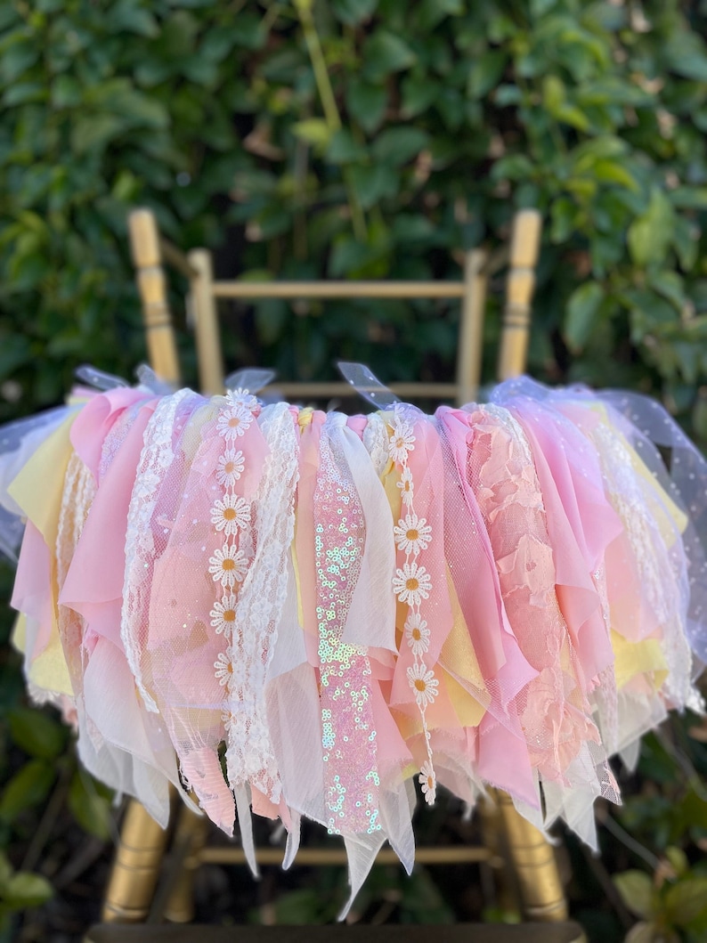 May include: A pink and yellow fabric garland with white daisy accents and a sparkly pink ribbon. The garland is hanging from a gold chair.